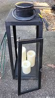 Black metal lantern with glass panels and three flameless LED candles inside. Front door of the lantern open revealing the candles.