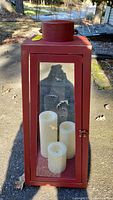 Front view of the red metal Candle Impressions lantern outdoors, showing three ivory flameless LED taper candles inside.
