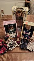 Eight porcelain dolls displayed with two still in original boxes on a wooden table, showing detailed clothing and hair styles.