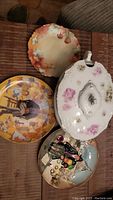Collector plates and soup tureen displayed on fabric surface, showing various artistic styles and floral porcelain tureen.