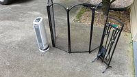 Full view of fireplace tool set on stand, fireplace screen, and tower space heater on outdoor concrete surface.