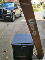 Dark-colored plastic hose hideaway storage box with lid, visible damage on one side, located on driveway.