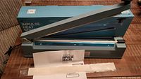 Impulse heat sealer machine in gray color with long handle and its box in the background on a fabric surface. A user manual and sealing strips are also placed in front of the unit.