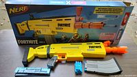 Front view of Nerf Fortnite AR-L motorized dart blaster with housing box visible in background.