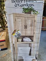 Photo of box with Better Homes & Gardens branding and product image showing the storage cabinet over toilet with plants and towels displayed.