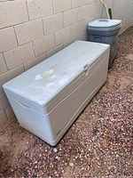 Full view of rectangular large outdoor storage bin placed on rocky ground against a wall, showing top and front side with lid closed.