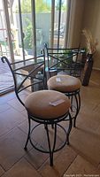 Pair of swivel bar stools with black metal frames and beige cloth cushioned seats, shown near a sliding glass door on tiled floor.