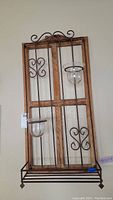 Wooden and metal rectangular hanging candle rack featuring decorative scrollwork and two glass candle holders attached, shown mounted on a wall.