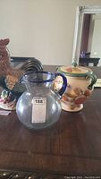Ceramic rooster figurine, glass pitcher with blue rim and ceramic biscotti pot with lid arranged on wood surface