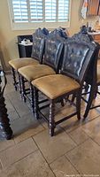 Five views showing three of six dark wood carved bar height dining chairs with tufted leather backs and cloth seats, ornate top backrest, and turned legs.