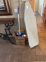 Photo of striped ironing board leaning against wall and box with cleaning supplies on tiled floor