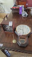 Photo showing both clocks on a wooden table: the wall clock with the pendulum detached and a smaller square desk clock with white face and markings.