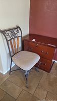 Photo showing metal chair with beige cushioned cloth seat and wood and scroll decorative backrest next to wood grain two drawer filing cabinet.