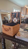 Woven wicker picnic basket open showing interior place settings with plates and cutlery and DaVinci Mandala game box on table.