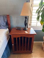 Full view of wood nightstand with ceramic lamp and clock on top beside a bed and window.