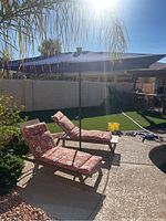 Blue poolside umbrella over two patterned cushioned lounge chairs near pool.
