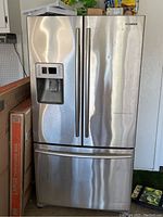 Front view of the Samsung refrigerator showing stainless steel French doors, water and ice dispenser with electronic display panel, and bottom freezer drawer.