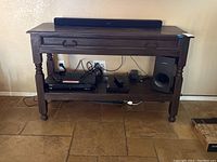 Solid wood side table shown with electronic devices on shelf and mounted TV above, showing top drawer, rectangular shape, turned legs.