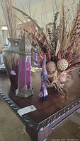 Photo showing pink tone glass candle holder, purple toned glass vase, copper tone pot with faux flowers, and painted ceramic vase with faux branches on a wooden table.