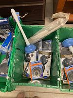 Photo showing a green bin containing four boxed showerheads, two packages of metal sink strainers, a white plunger with blue rubber end, and a wrapped safety grab bar.