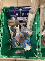 Green bin containing various plumbing and hardware supplies including showerheads, a faucet box, strainers, a plunger and safety bar.