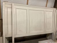 Wide shot of large king size white headboard leaning against a wall in a storage area. Shows five panel design with simple molding detail. Surface shows dirt and discoloration.