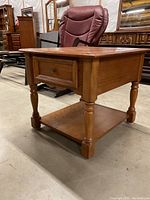 Full view of oak end table showing the turned legs, drawer with metal knob, and lower shelf, along with visible wear and scratches.