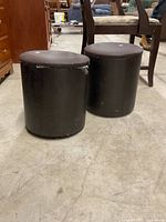 Two black cylindrical ottoman stools side by side. One stool shows visible wear with scuffs and peeling on the upholstery. Both stools approximately 13 inches tall and 12 inches in diameter. The stools are placed on a concrete floor with wooden furniture in the background.
