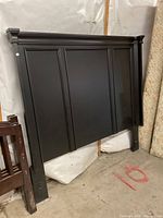 Photo shows a black wooden queen-size headboard with three vertical panels and decorative molding at the top, positioned against a wall with surrounding furniture items visible.