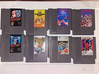 Photo of 8 NES game cartridges laid out on a white surface showing front labels: Super Mario Bros., Caveman Games, Tetris, Dr. Mario, Hogan's Alley, Bionic Commando, Marble Madness, The Karate Kid.