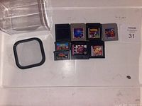 Top-down photo showing eight retro game cartridges arranged on a flat surface next to a clear plastic container lid.