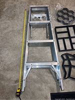 Full view of 4 step aluminum ladder lying on the floor next to a yellow measuring tape showing its length. Includes black cement moulding frames nearby.
