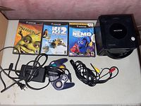 Nintendo GameCube console, power adapter, AV cables, one controller and three game cases (Shrek 2, Ice Age 2: The Meltdown, Finding Nemo) on table
