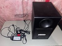 Photo of Samsung subwoofer with power cable and adapter on a table against a pink insulation background.