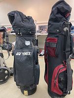 Side view of two golf bags, one branded Yonex with visible logo and multiple zippered compartments, next to another black and red golf bag.