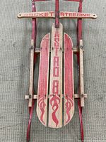 Top view of the sled showing the wooden plank labeled 'ASTRO 8' with flame designs and the wooden 'ROCKET STEERING' handle bar.