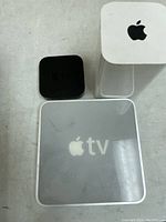 Photo showing two Apple TV devices, one black matte box and one white translucent topped box with Apple logo.