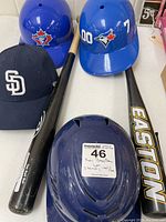 Photo showing two blue Blue Jays batting helmets, two Easton baseball bats, a navy San Diego Padres New Era hat, and a navy batting helmet with ventilation slots.