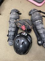 Wilson knee and shin guards, Rawlings catcher's glove, and a black helmet displayed on carpeted floor.