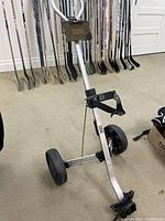 Side angle showing the aluminum frame, two large black rubber wheels, securing straps, and foldable design of the golf bag carrier among golf clubs.