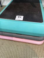 Stacked fitness steps in teal, black and pink on carpet floor, showing rubber decking surface and rounded edges.