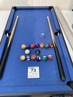 Mini blue felt pool table with two wooden cues, 16 balls arranged in two rows, and blue chalk on the table