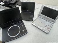 Photo showing three portable DVD players including brands Polaroid and Phillips, all with foldable screens and circular DVD trays, in black and silver.