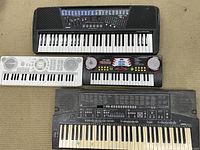 Lot of four electronic keyboards varying in size and color arranged on carpet. Includes one large Casio keyboard, a small white keyboard, a black keyboard with red and white design, and an older heavily worn keyboard with missing keys.
