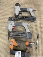 Three pneumatic nail tools - two Porter Cable and one Rigid - laid on carpeted floor with pen for scale