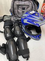 Six Six One black knee and shin guards with protective plastic plates and adjustable straps next to Roots bag in background