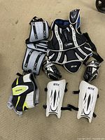 STX hockey protective gear including shoulder pads, arm guards, shin guards in black and white colors alongside Brine Maximus gloves in black, white, neon green, and gray.