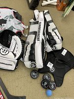 Top-down photo showing Warrior kids goalie leg pads, catch glove, blocker, four black pucks and a blue foam ball on a carpet.