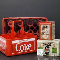 Photo showing red plastic Coke crate and family pack carrier alongside two vintage Coca-Cola collectible tins with decorative artwork.