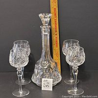 Photo of crystal decanter with stopper set in center, surrounded by four matching wine glasses. Wooden ruler placed at back for size reference.